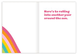 The Roller Skates Birthday Card by Lauren-Ashley Barnes features an open illustration with bold rainbow stripes in the corner and “Here’s to rolling into another year around the sun.” in pink bubbly text on the right page.