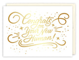 The New Human New Baby Card by Andrea Williams features gold cursive text—“Congrats on Your New Human!”—with decorative swirls, dots, and stars. Printed in the USA on recycled paper; envelope partially shown behind the card.