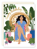 The Plant Queen Birthday Card by Lauren-Ashley Barnes features a woman with long dark hair in a blue polka-dot dress on a rattan chair, surrounded by potted plants, with “Happy Birthday Plant Queen” in gold text.