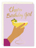 The Cheers Birthday Girl Card by Lauren-Ashley Barnes features a pink background, a hand holding a yellow cocktail with lime, and gold script that says Cheers Birthday Girl. Comes with an envelope made from recycled paper.