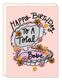 The Total Babe Birthday Card by Natalie Henry Charles features a pink background, an illustrated clam shell with flowers, recycled paper, and hand-lettered text reading “Happy Birthday” and “To a Total Babe.”.