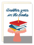 The In The Books Birthday Card by Tiffany Grimes features a stack of books on a pink cake stand topped with a lit candle and blue script text that reads, Another year in the books. Perfect for any bookworm’s birthday.