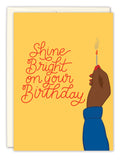 The Shine Bright Birthday Card by Tiffany Grimes features “Shine Bright on your Birthday” in red script, a dark-skinned hand with red nails holding a lit match on a yellow background, and is printed on recycled paper.