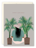 The Growth Birthday Card by Tiffany McGraw features a person with long black hair and a green shirt, seated between two potted palms on a gray background. HAPPY BIRTHDAY! is printed on recycled paper, symbolizing growth.