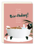 The Soak In Love Birthday Card by Tiffany McGraw showcases a woman enjoying a bubble bath with a cupcake and candle, “SOAK” and “Birthday!” in the background, potted plants around the tub, and is printed on recycled paper.